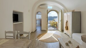 A room with a TV, a dining area with chairs and a table, a wardrobe, and a view of the sea. - Just Blue (Santorini)