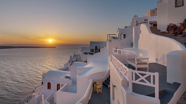 A coastal building with white architecture, a staircase, and a view of the sea at sunset. - Just Blue (Santorini)