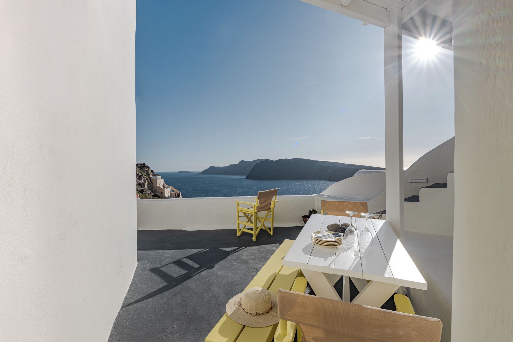A terrace with a white table and yellow chairs, overlooking a coastal landscape with cliffs and the sea.