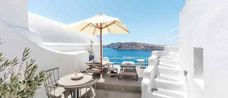Terraço de paredes brancas com mesa e cadeiras, um guarda-sol bege e vista para o mar e terra distante.