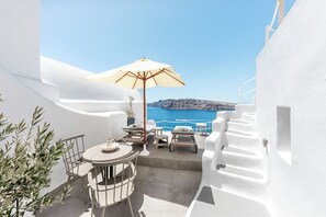A white-walled terrace with a table and chairs, a beige umbrella, and a view of the sea and distant land. - Just Blue (Santorini)
