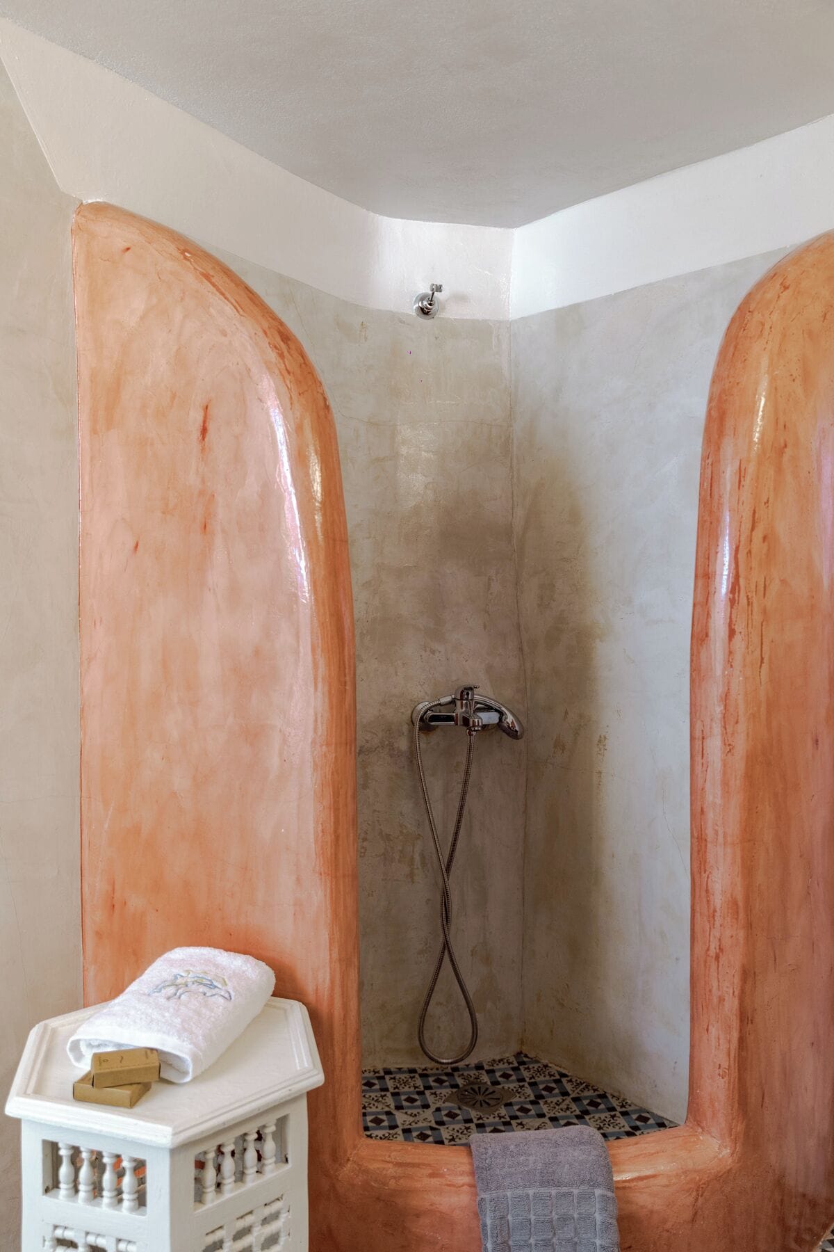 A shower with a handheld showerhead, a towel on a shelf, and a patterned floor.