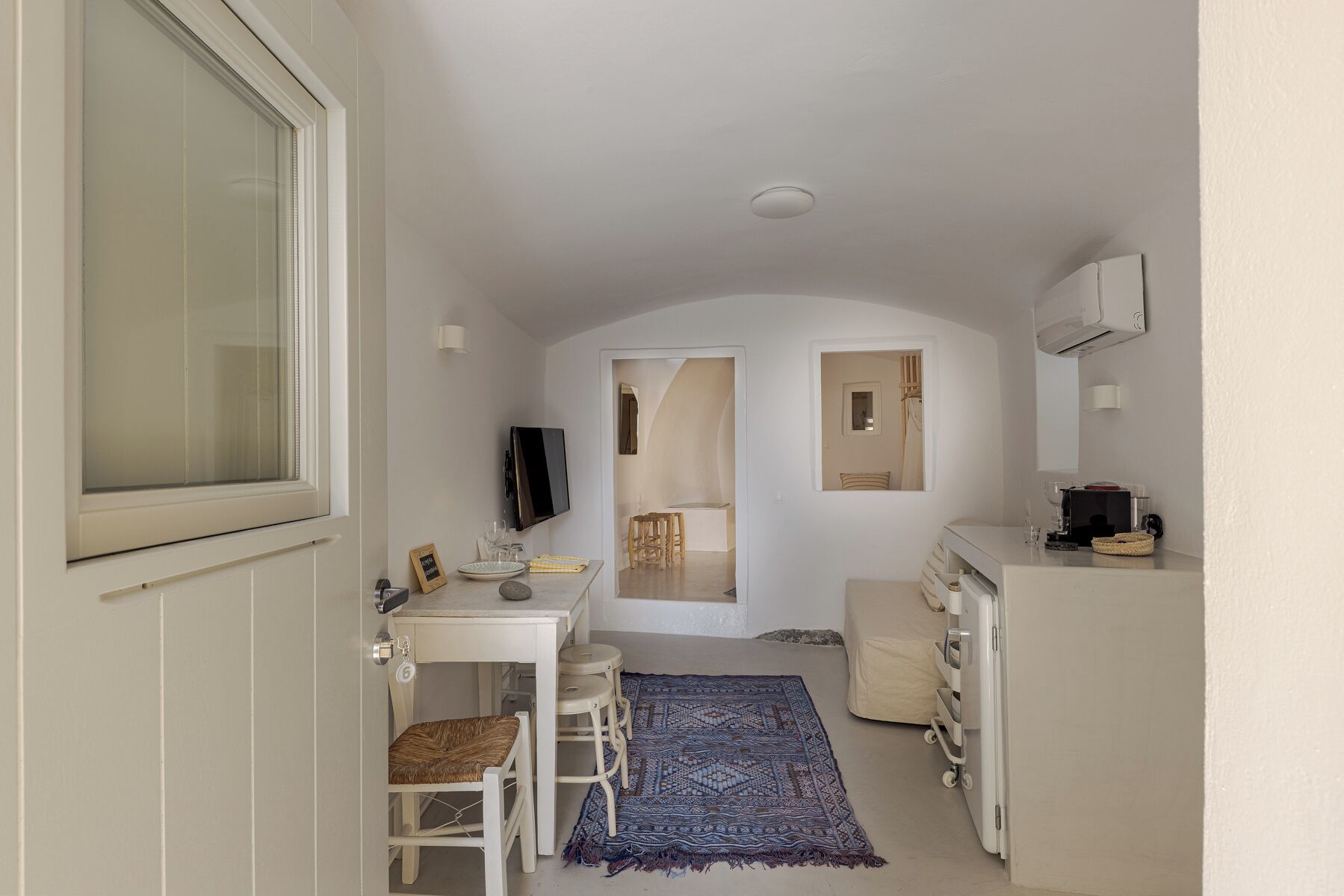 A narrow, white-walled room with a small dining table, chairs, a TV, and a kitchenette area.