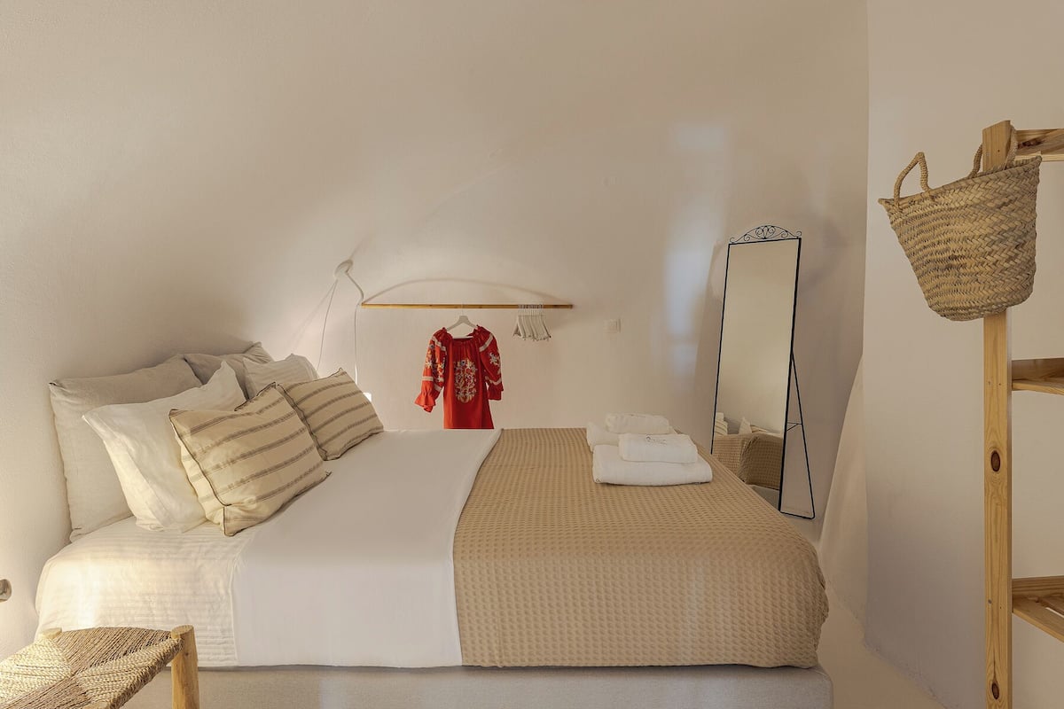 Un dormitorio con una cama, almohadas, una prenda roja colgada, un espejo y una cesta tejida.