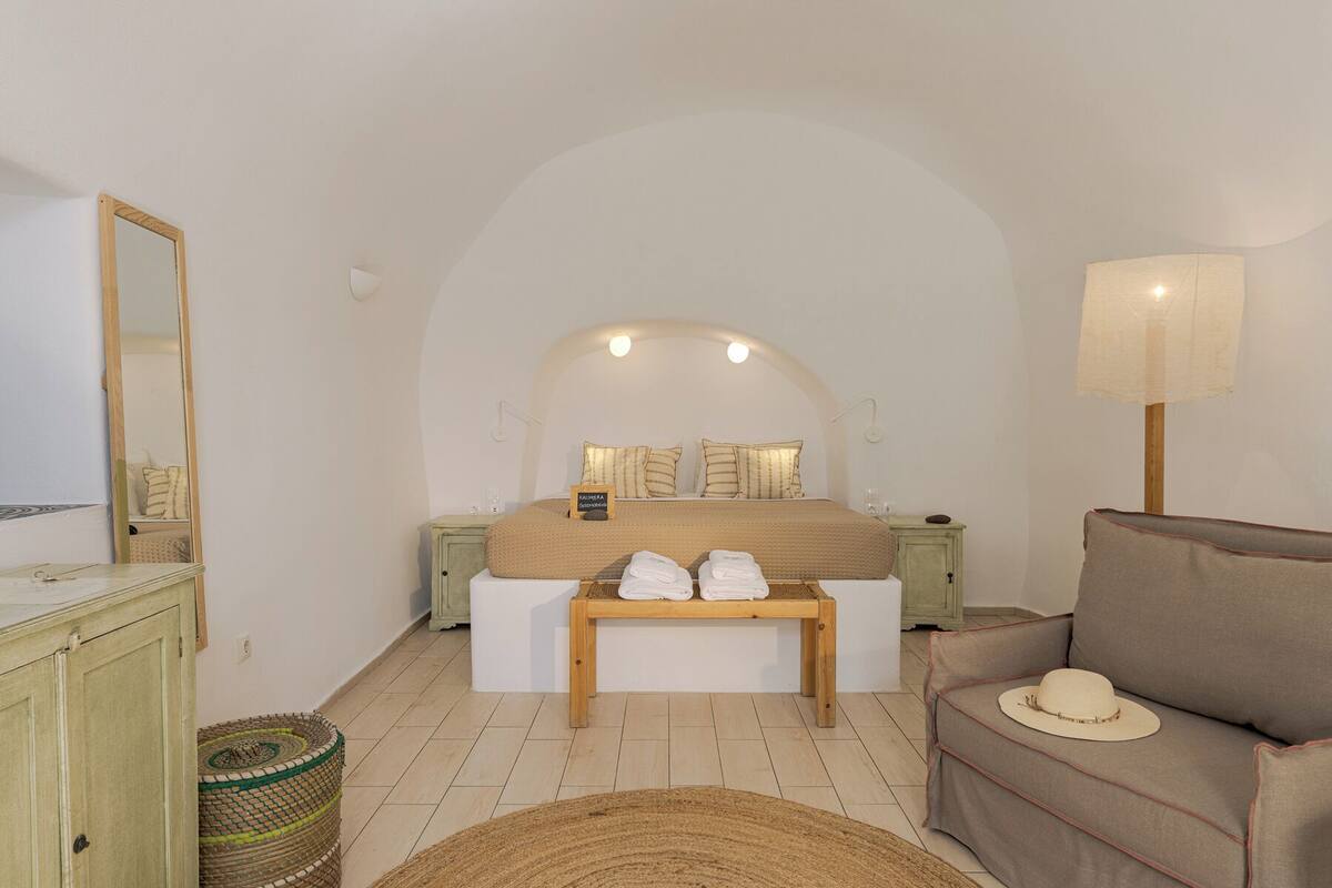 Un dormitorio con cama, mesitas de noche, un banco, un sofá, una lámpara, un espejo y una cesta de mimbre.