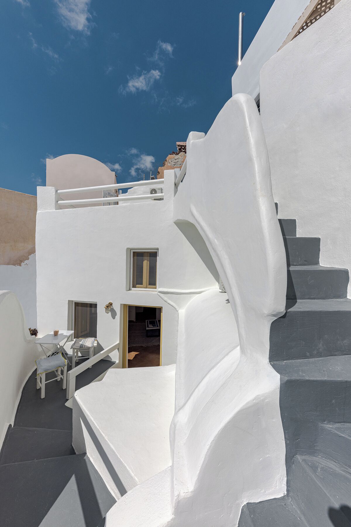 Un edificio blanco con una escalera que lleva a una puerta, una mesa pequeña con una silla y una ventana.
