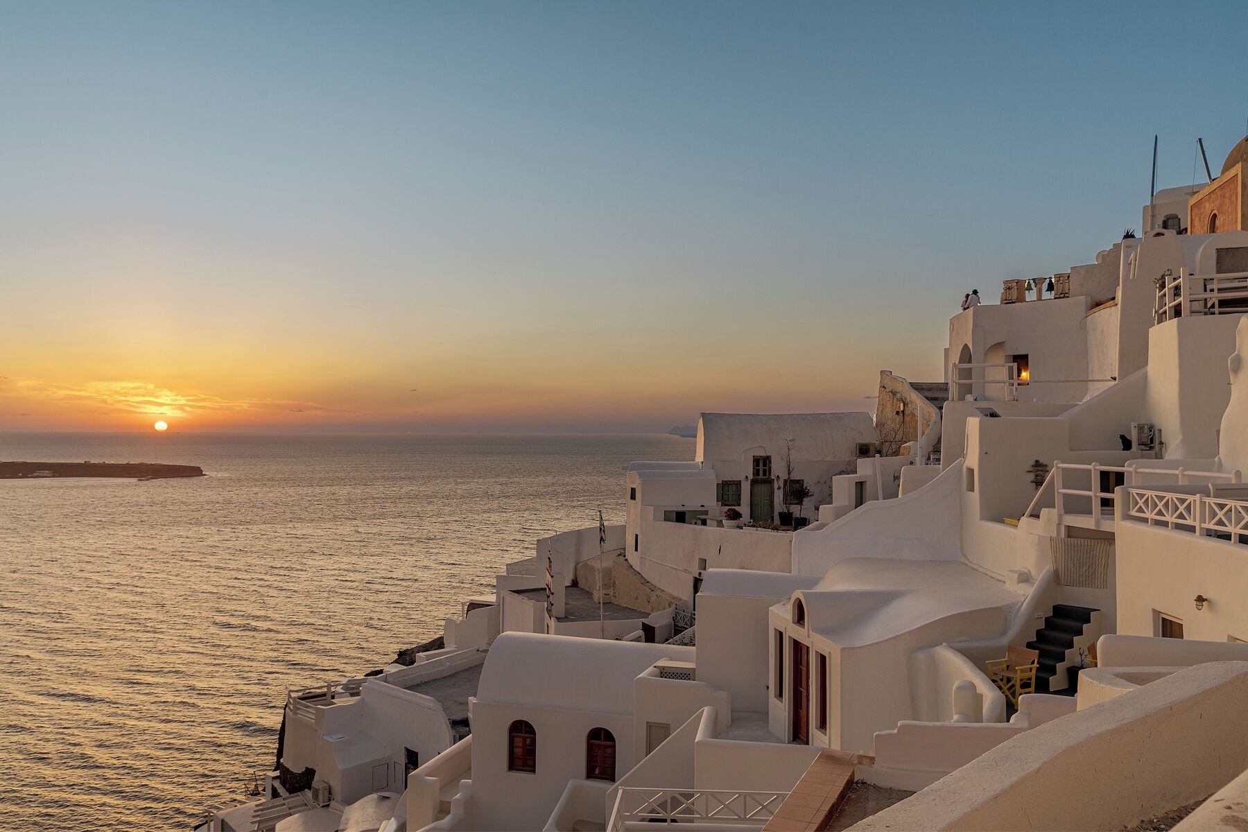 Un pueblo costero con edificios blancos, cielo despejado y el sol poniéndose sobre el mar.