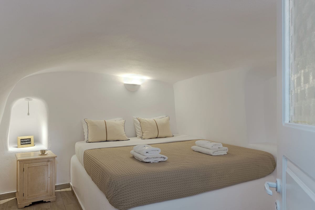 Un dormitorio con cama, almohadas, una mesita de noche y una lámpara de pared.