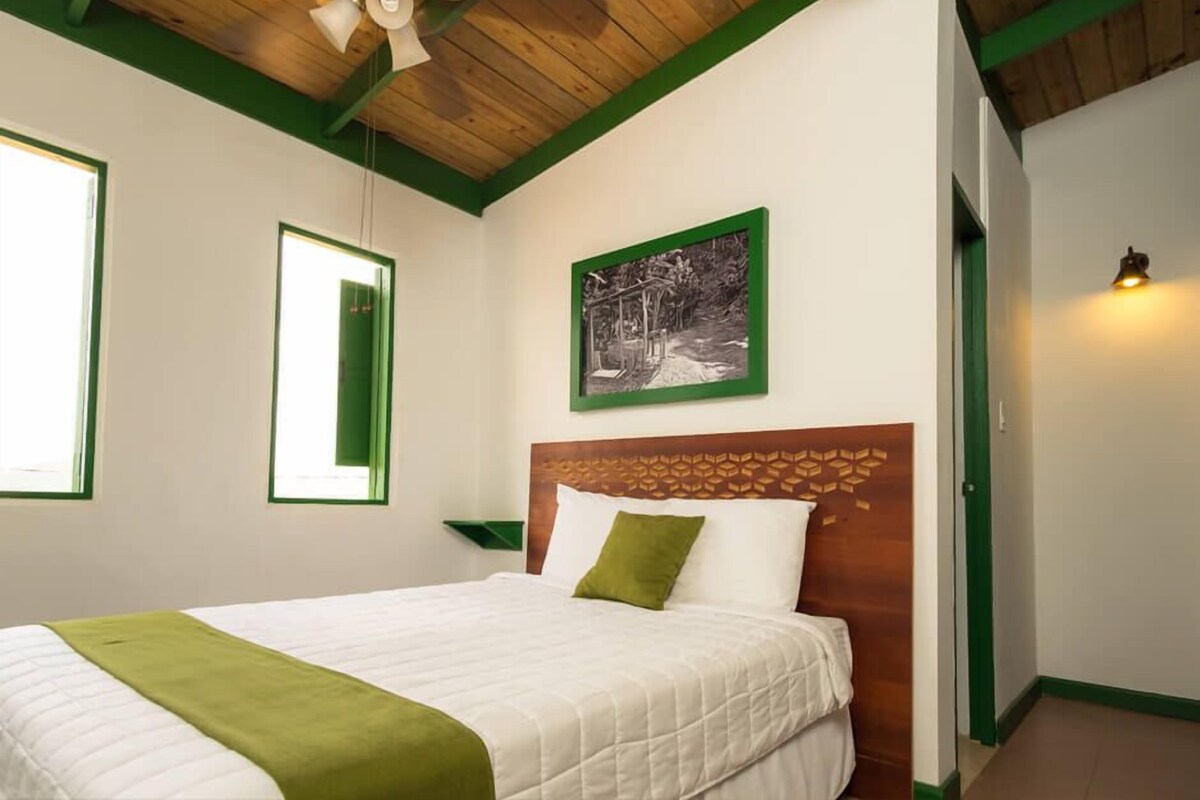 A bedroom with a wooden headboard, a bed with white bedding and a green throw, a framed black and white photo on the wall, and a green-framed window.
