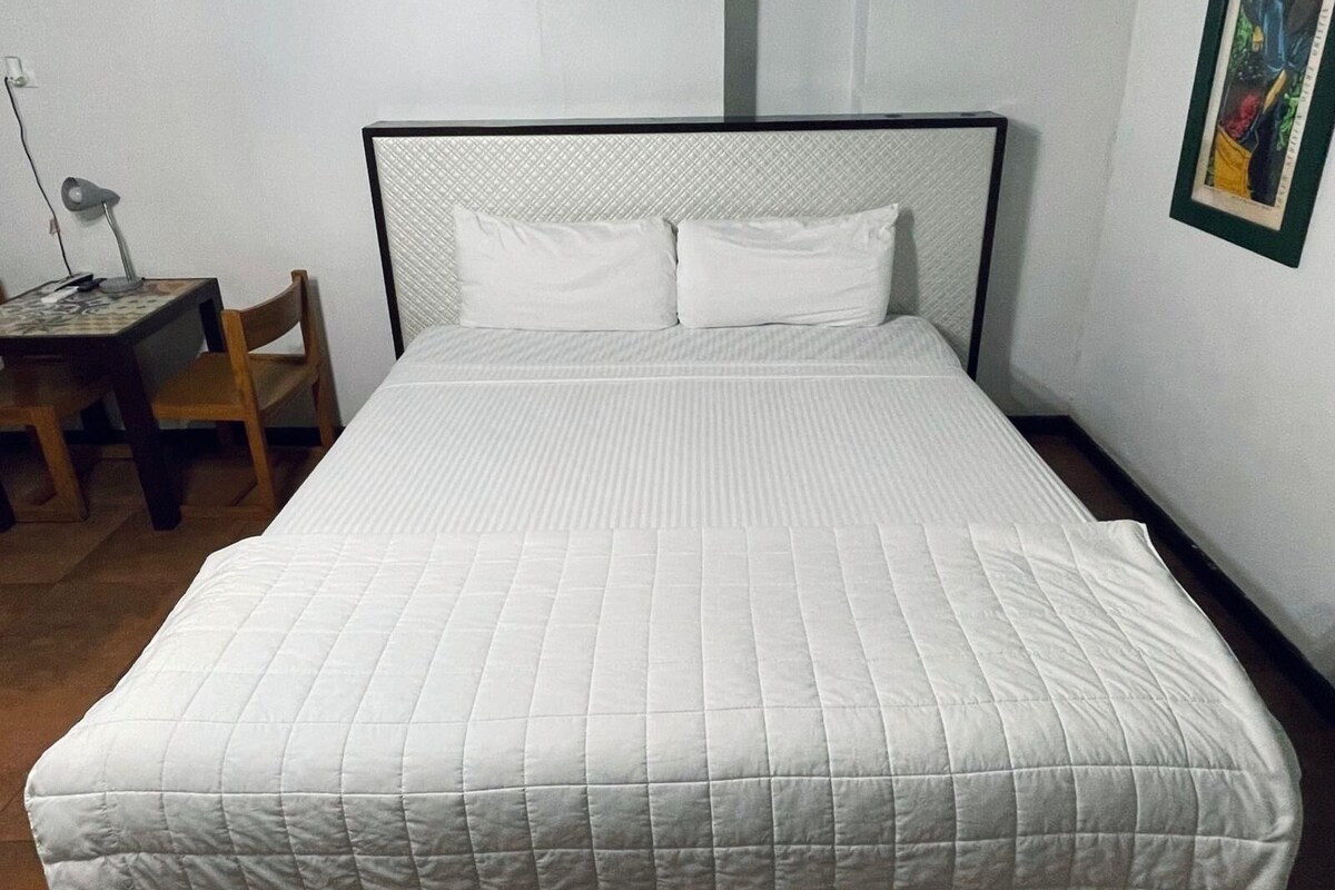 A neatly made bed with a white quilted bedspread and two pillows, a wooden chair, and a desk with a lamp.