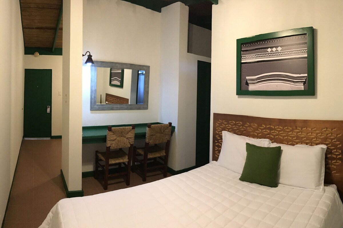 A hotel room with a bed, a desk with two chairs, a mirror, and a wall-mounted picture.