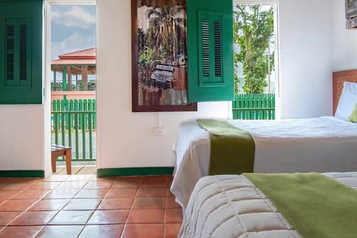 A bedroom with a bed, a window with a view of a green fence and trees, and a painting on the wall.