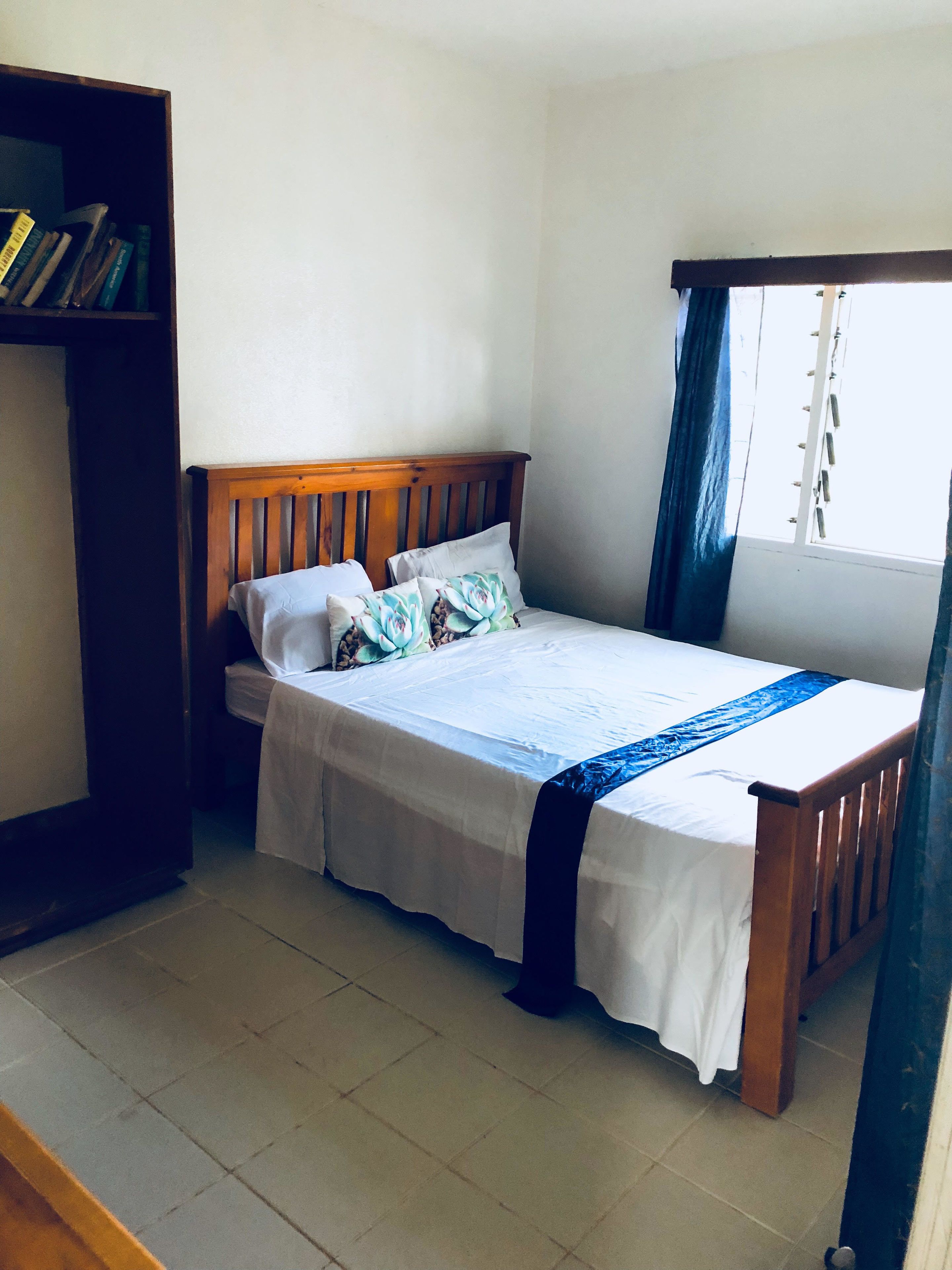 A single bed with a wooden headboard, white bedding, and a blue runner. There is a bookshelf to the left and a window with curtains to the right.