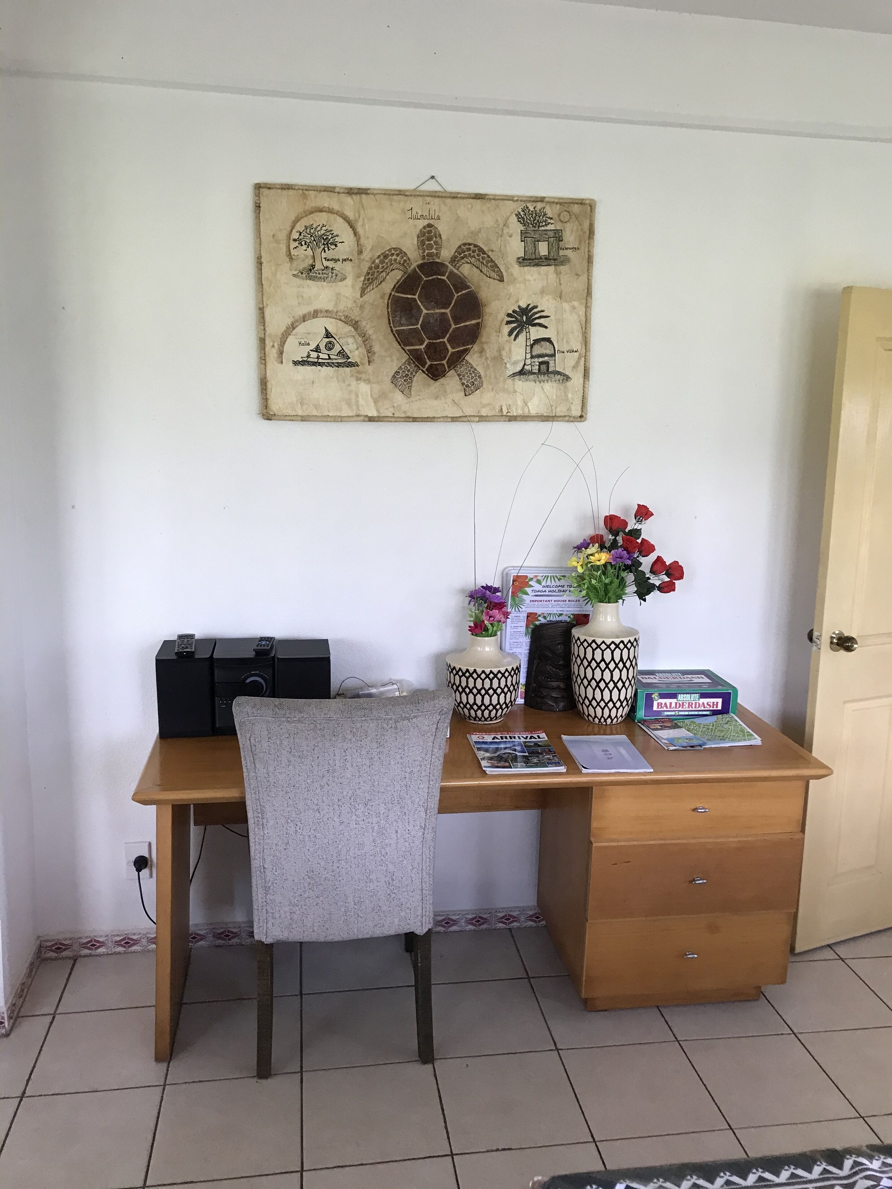 A wooden desk with a chair, a wall-mounted artwork depicting a turtle and palm trees, a vase with flowers, and a Balderdash game box.