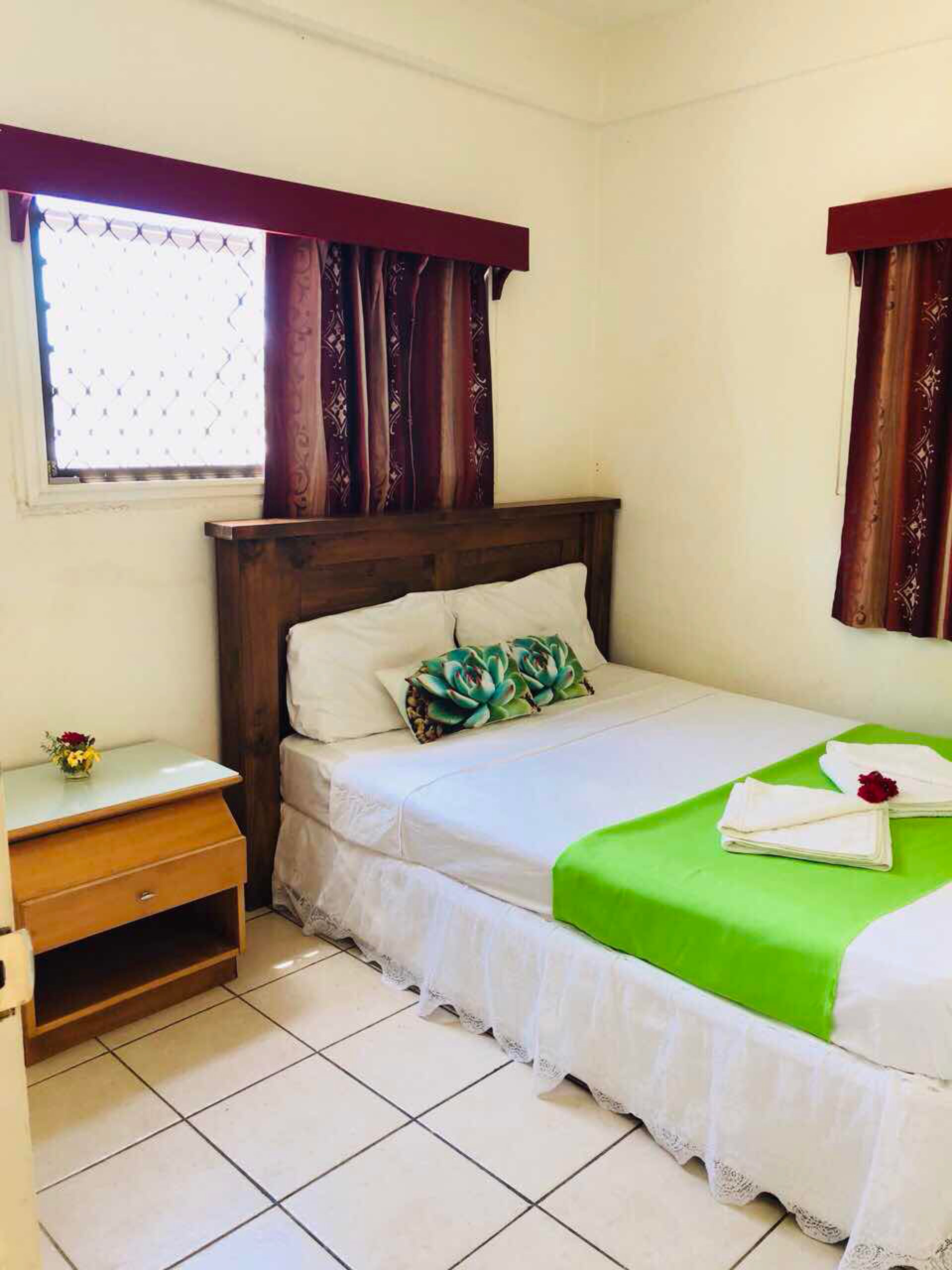 A hotel room with a bed, a nightstand, a window with curtains, and a small table with a vase of flowers.