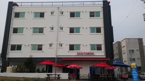 Un edificio de varios pisos con el nombre 'SANTORINI' en la fachada. Cuenta con espacios de estar al aire libre con sombrillas rojas, y un letrero que indica que es una casa de huéspedes.
