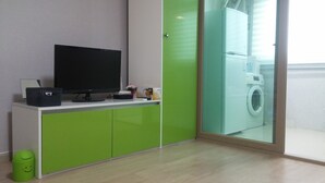 Una habitación moderna con una televisión de pantalla plana sobre un mueble blanco, una lavadora visible a través de una puerta de vidrio y un bote de basura verde con una carita sonriente.