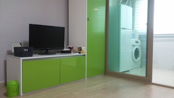 Una habitación moderna con una televisión de pantalla plana sobre un mueble blanco, una lavadora visible a través de una puerta de vidrio y un bote de basura verde con una carita sonriente.