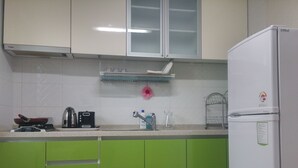 Una cocina con gabinetes verdes, un refrigerador blanco, una estufa a gas, una tetera, un tostador, un escurreplatos y una flor rosa en la pared.