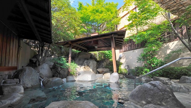 Un onsen japonés tradicional con un pasillo de madera sobre un estanque rocoso, rodeado de vegetación y una estructura de bambú.