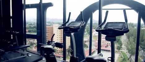 Un gimnasio con bicicletas estáticas y una cinta de correr, con amplias ventanas que ofrecen vista a la ciudad.