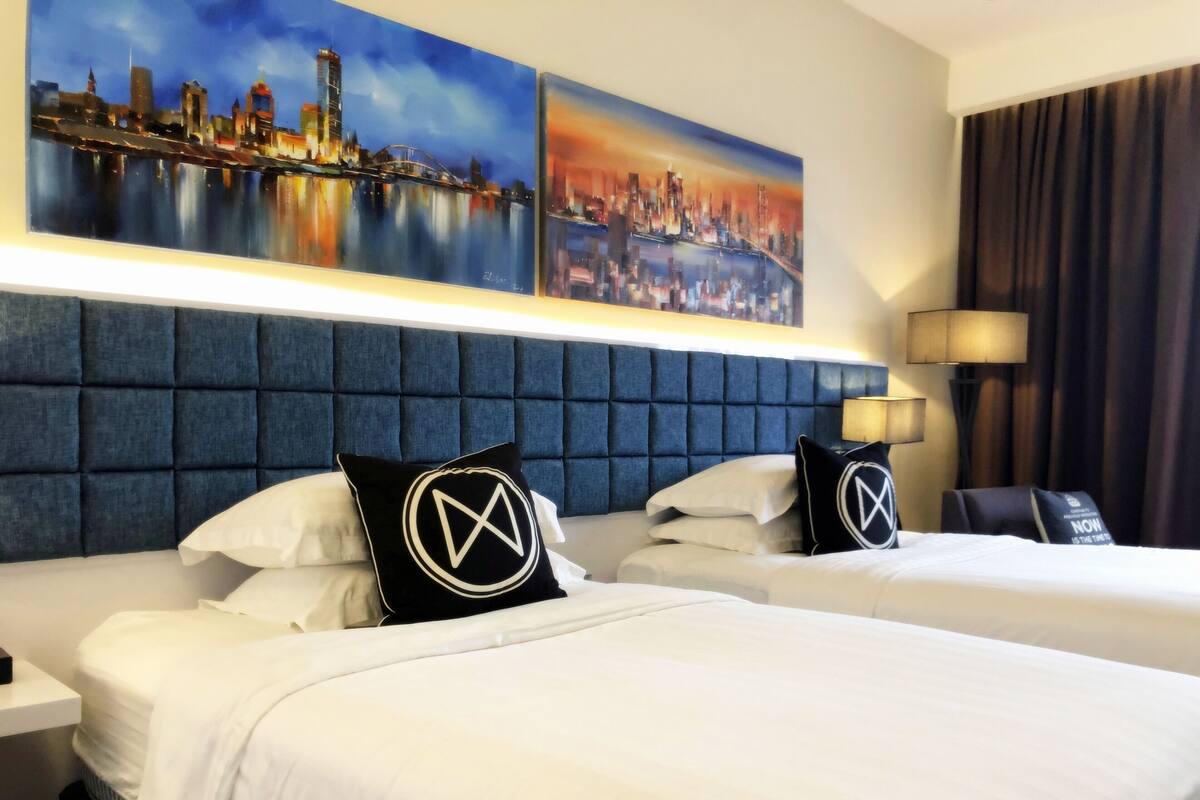 Een hotelkamer met twee bedden, een blauw hoofdbord, witte beddengoed en twee schilderijen van stadsgezichten aan de muur.