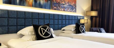 A hotel room with two beds, a blue headboard, white bedding, and two paintings of cityscapes on the wall.