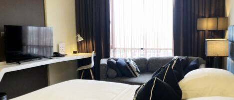 A hotel room with a bed, a desk with a TV, a chair, a sofa, and a window with curtains.