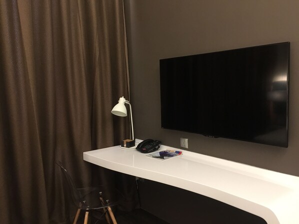 Una habitación de hotel moderna con televisión de pantalla plana montada en la pared, un escritorio blanco con lámpara, teléfono y una revista.