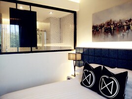 Una habitación de hotel con una cama grande, cabecero, mesita de noche, lámpara, espejo y un cuadro de la ciudad en la pared.