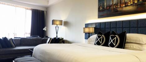 A hotel room with a large bed, a sofa, a bench, a painting of a cityscape, and two lamps.
