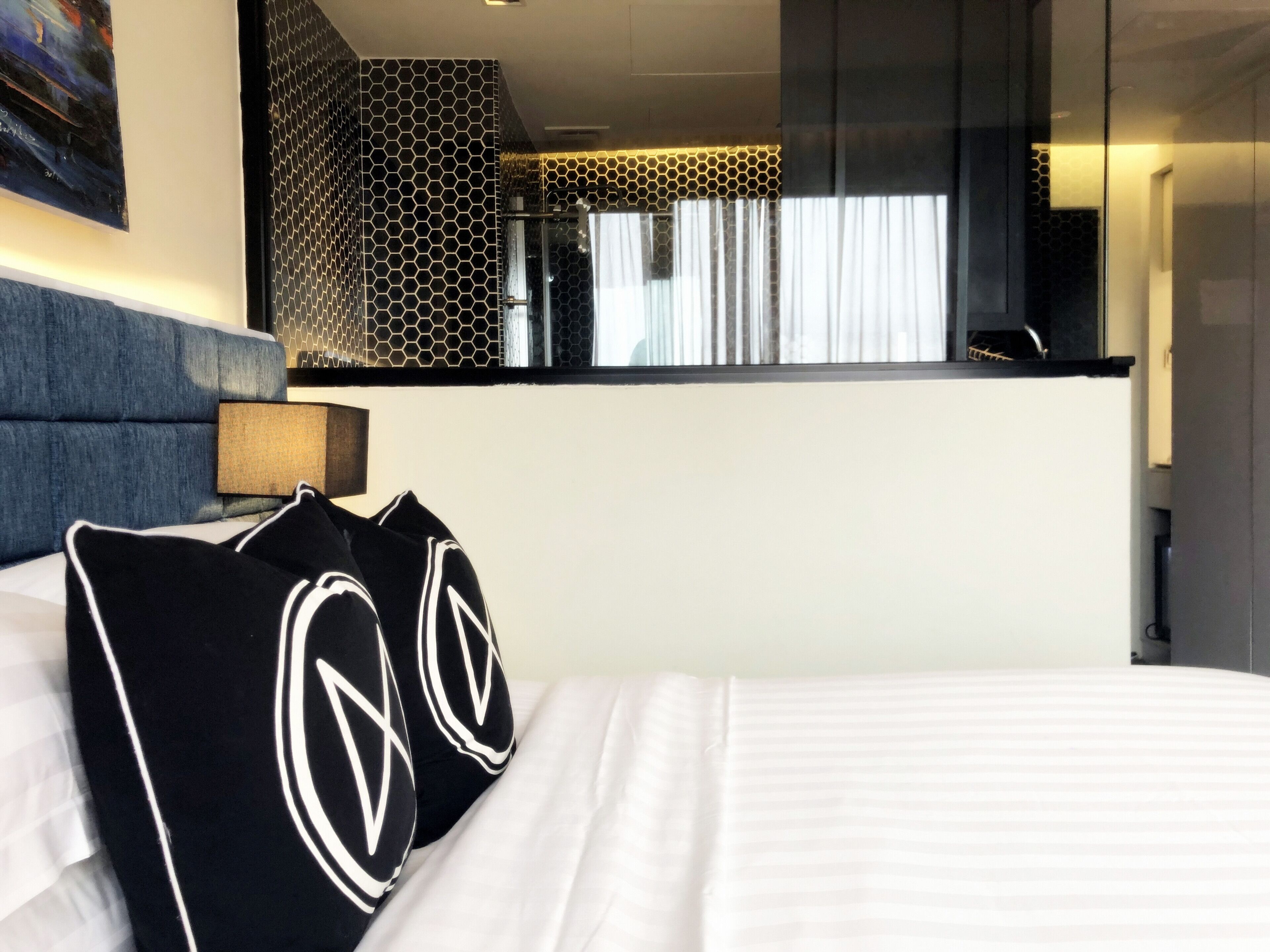 Una habitación de hotel con cama, dos almohadas con un logotipo, cabecera, mesita de noche, lámpara, ventana con cortinas y vista a un paisaje urbano.