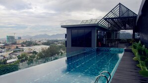 Ein Pool mit Endlosüberlauf auf dem Dach, mit einer Holzterrasse, einer überdachten Sitzgruppe und Blick auf die Stadt und die Berge im Hintergrund.
