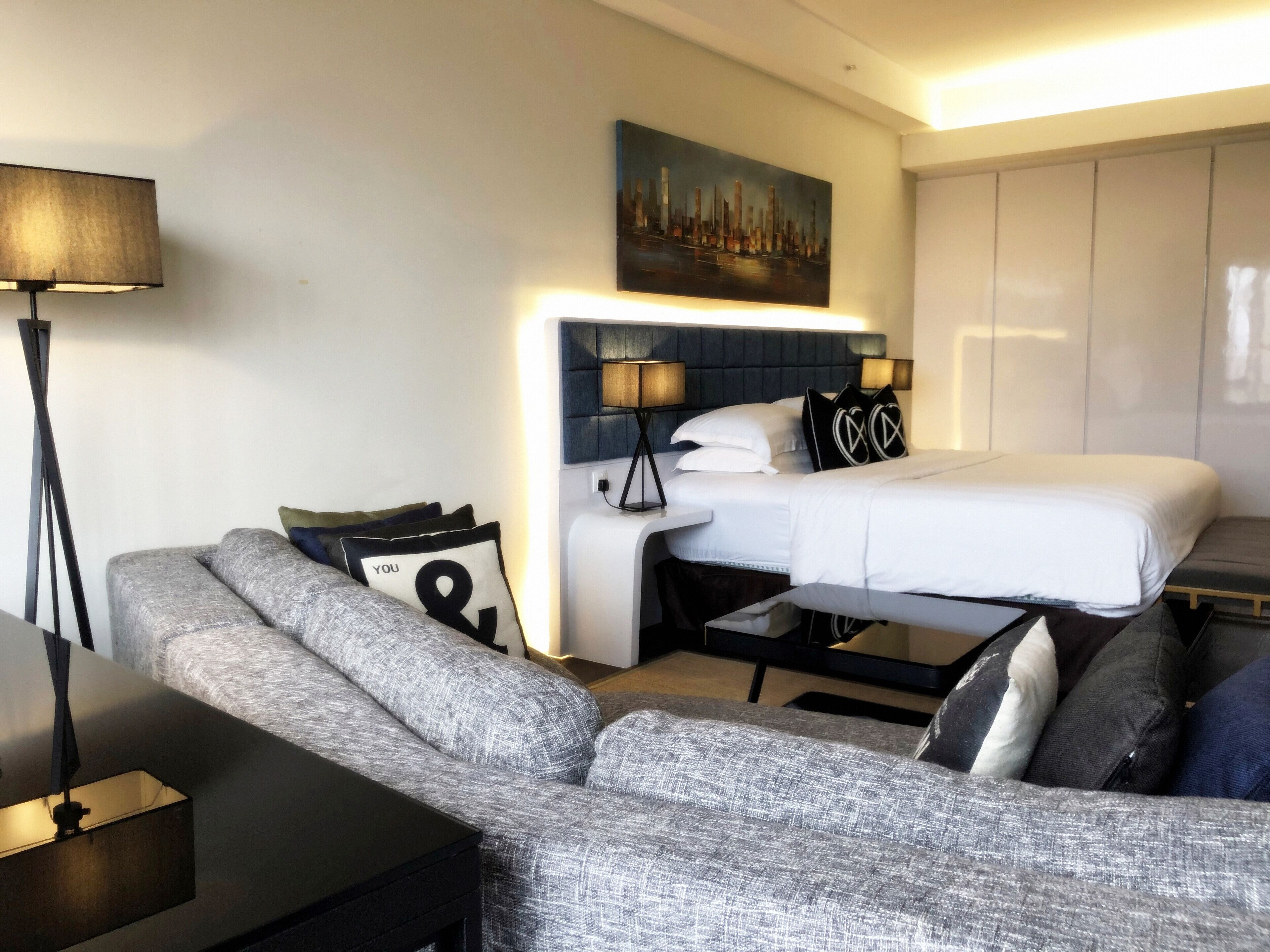 Una habitación de hotel moderna con un sofá gris, una cama con cabecera, una mesita de noche, una lámpara y un cuadro en la pared.