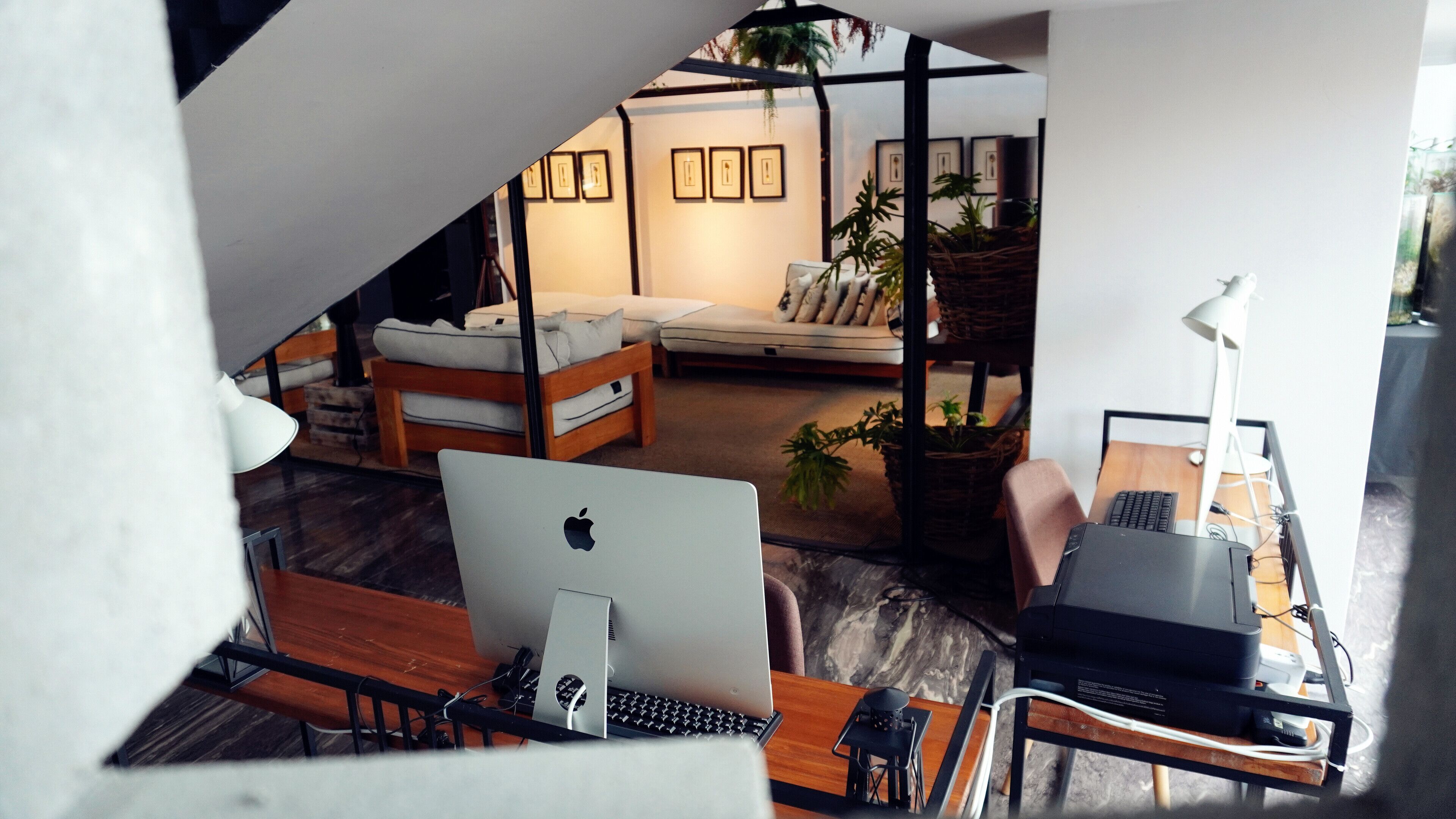A modern living room with a sofa, a desk with a computer, and a coffee table. There are framed pictures on the wall and a plant in a basket.