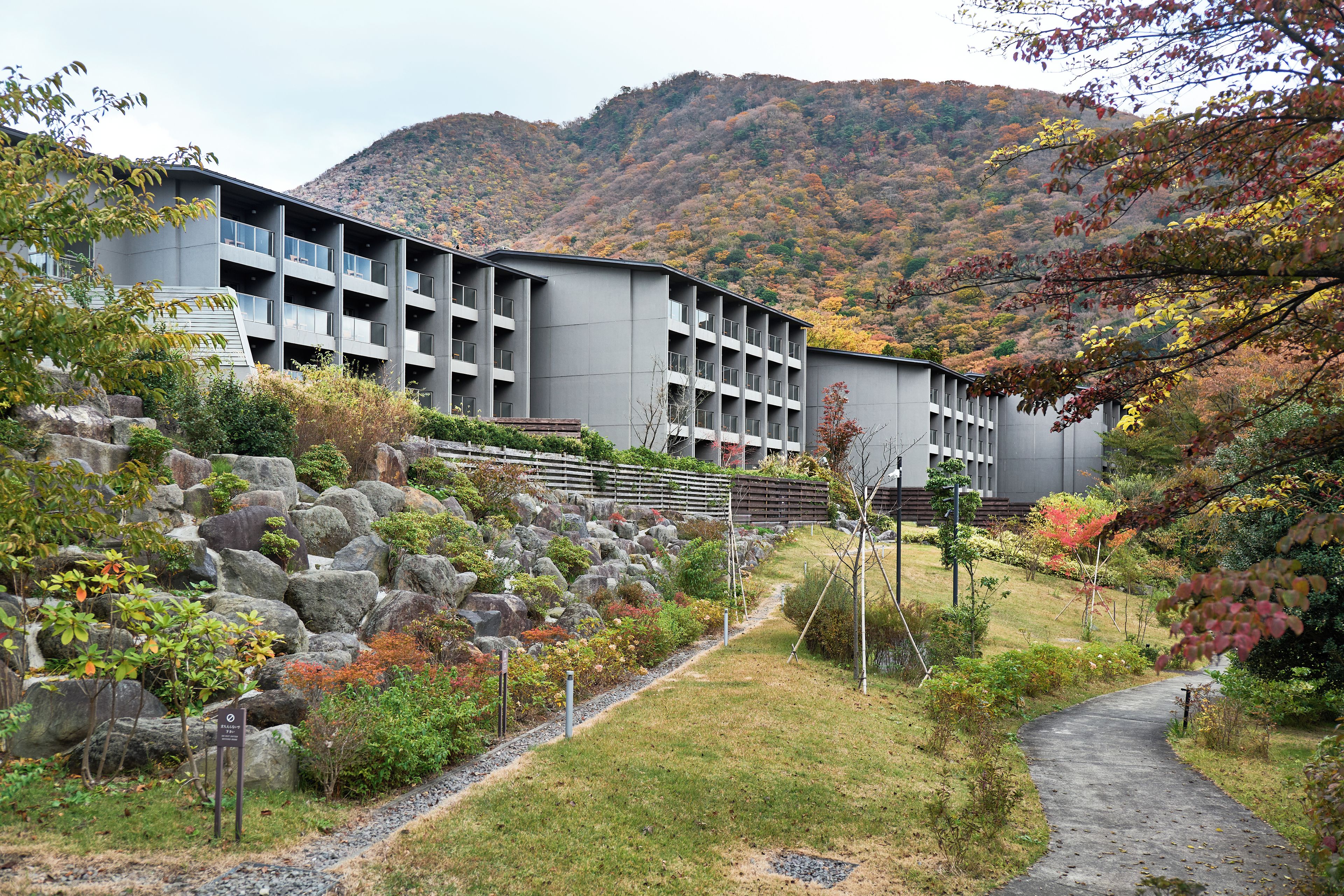 A modern building with multiple balconies, surrounded by a landscaped garden with rocks and shrubs, and a mountain in the background.