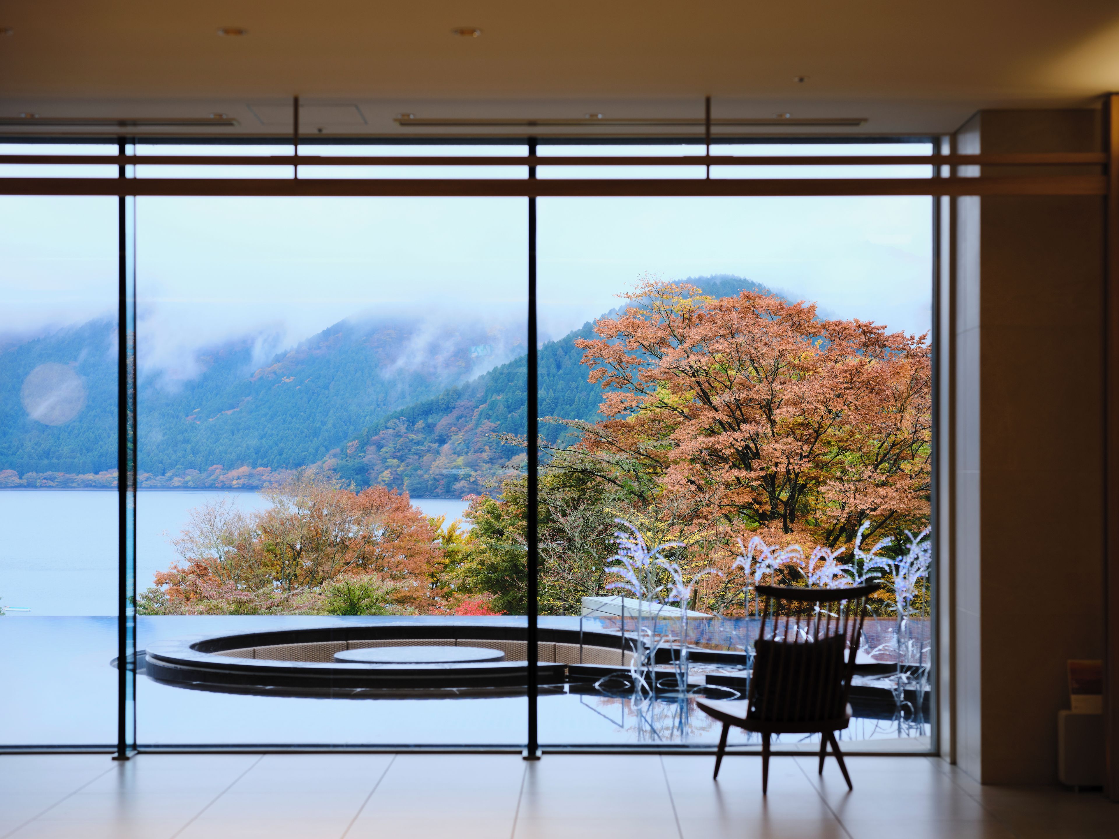 A room with a large window offering a view of a lake and autumn foliage.