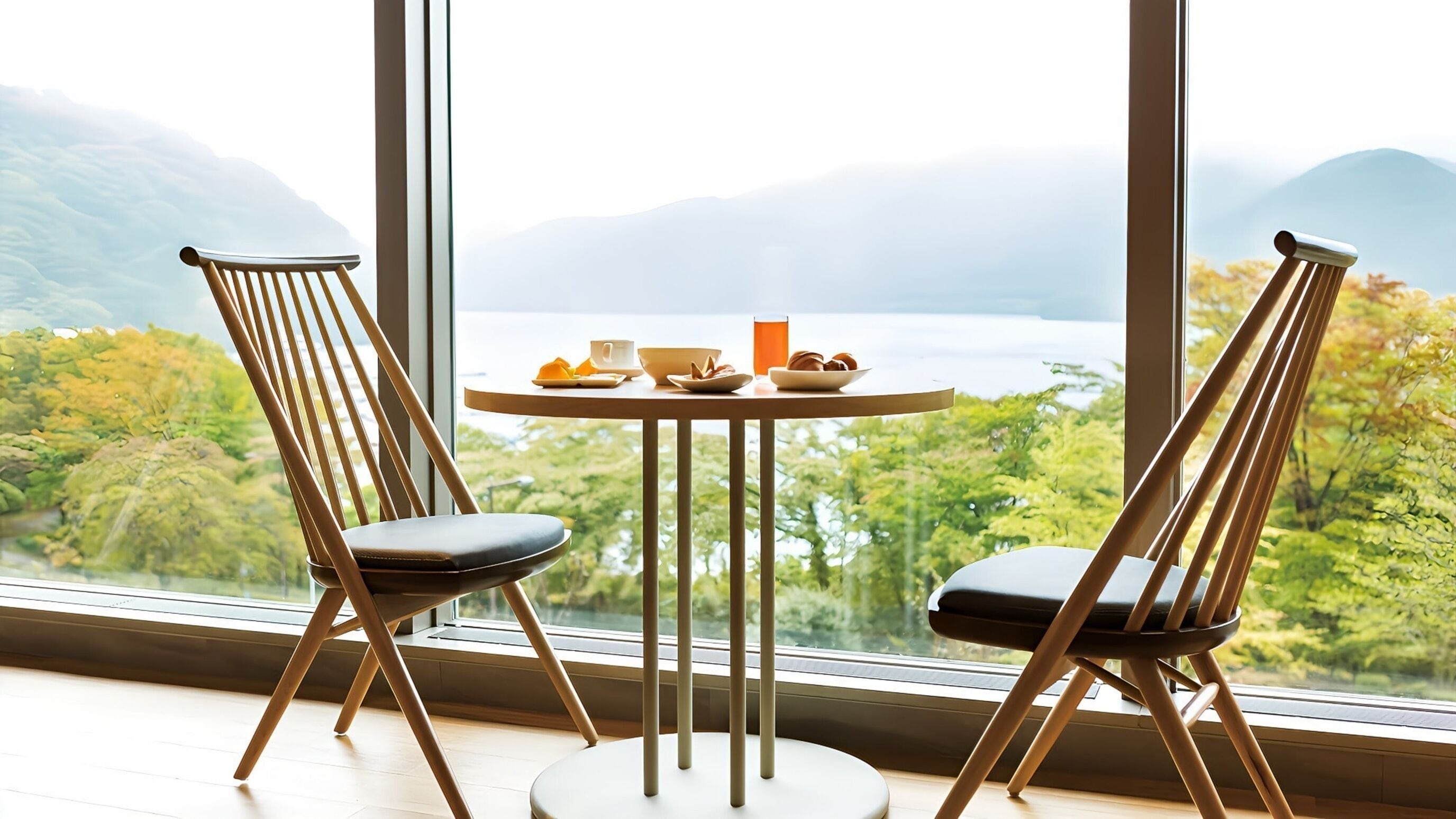 A dining area with two chairs and a table, a glass of orange juice, a bowl of fruit, and a cup, with a view of a lake and mountains through large windows.