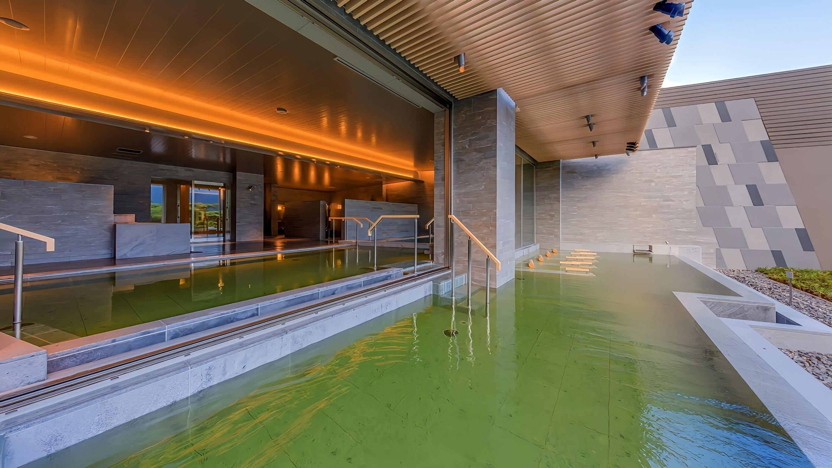 A modern indoor pool with a glass barrier, wooden steps, and a stone wall.