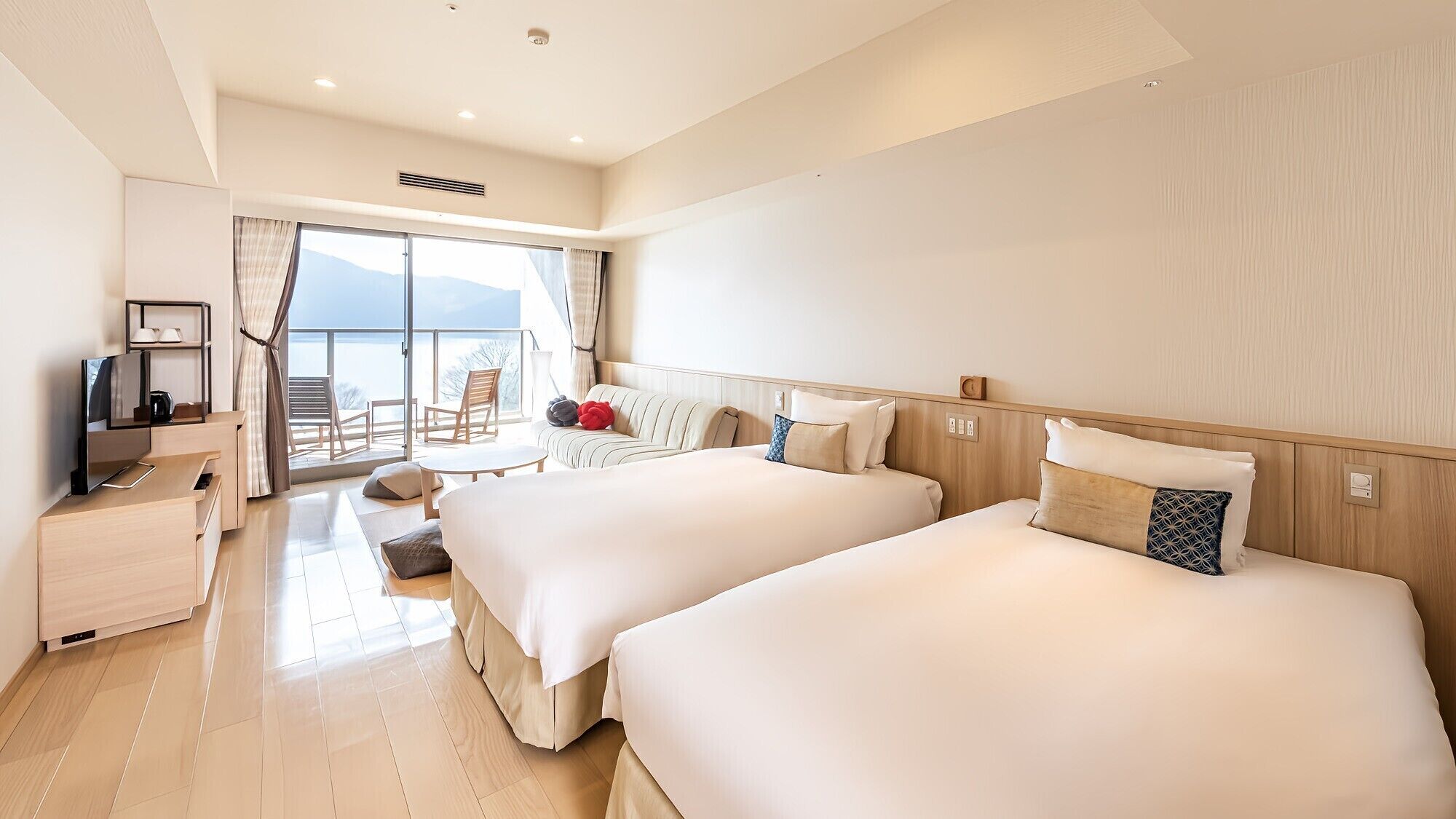 Ein Hotelzimmer mit zwei Betten, einem Schreibtisch mit Fernseher, einem Balkon mit Sitzgelegenheiten und einem großen Fenster.