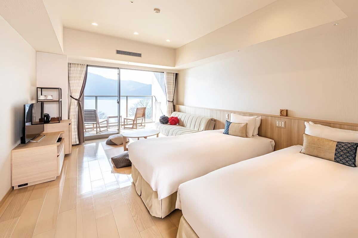 Ein Hotelzimmer mit zwei Betten, einem Schreibtisch mit Fernseher, einem Balkon mit Sitzgelegenheiten und einem großen Fenster.