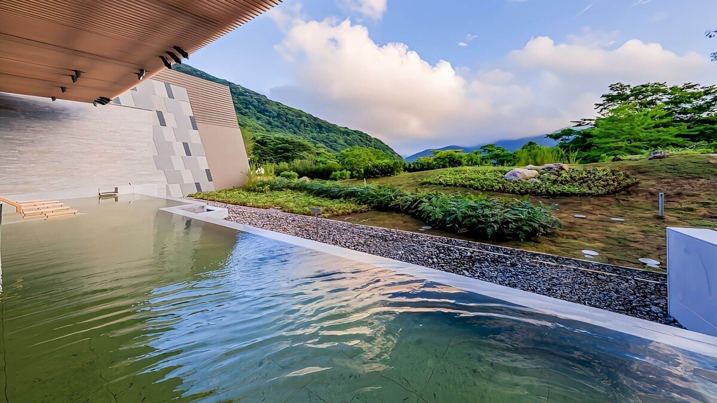 A modern building with a pool, surrounded by greenery and hills.
