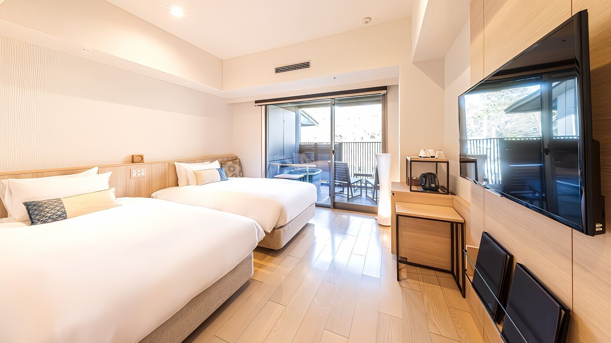 Ein Hotelzimmer mit zwei Betten, einem Fernseher, einem Balkon mit Tisch und Stühlen sowie einer Glastür.