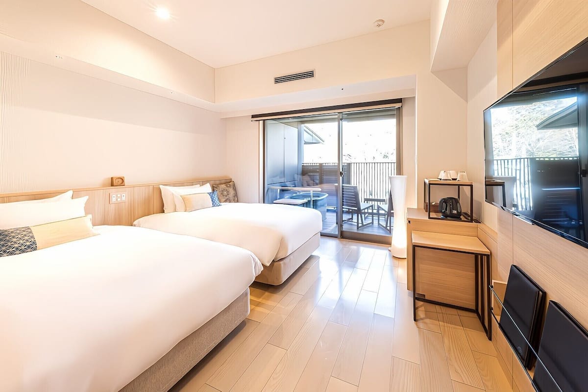 Ein Hotelzimmer mit zwei Betten, einem Fernseher, einem Balkon mit Tisch und Stühlen sowie einer Glastür.