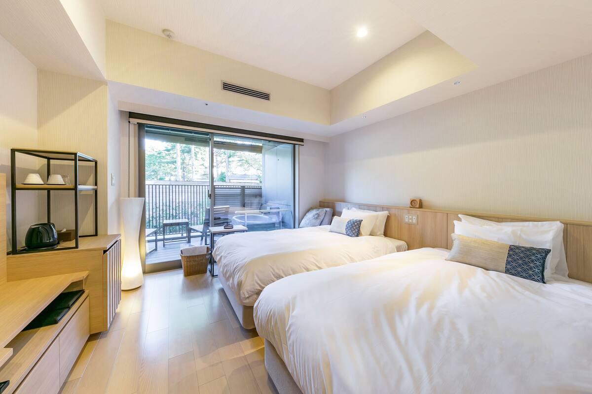 Ein Hotelzimmer mit zwei Betten, einem Schreibtisch, einem Stuhl, einem großen Fenster und einem Balkon mit Gartenmöbeln.