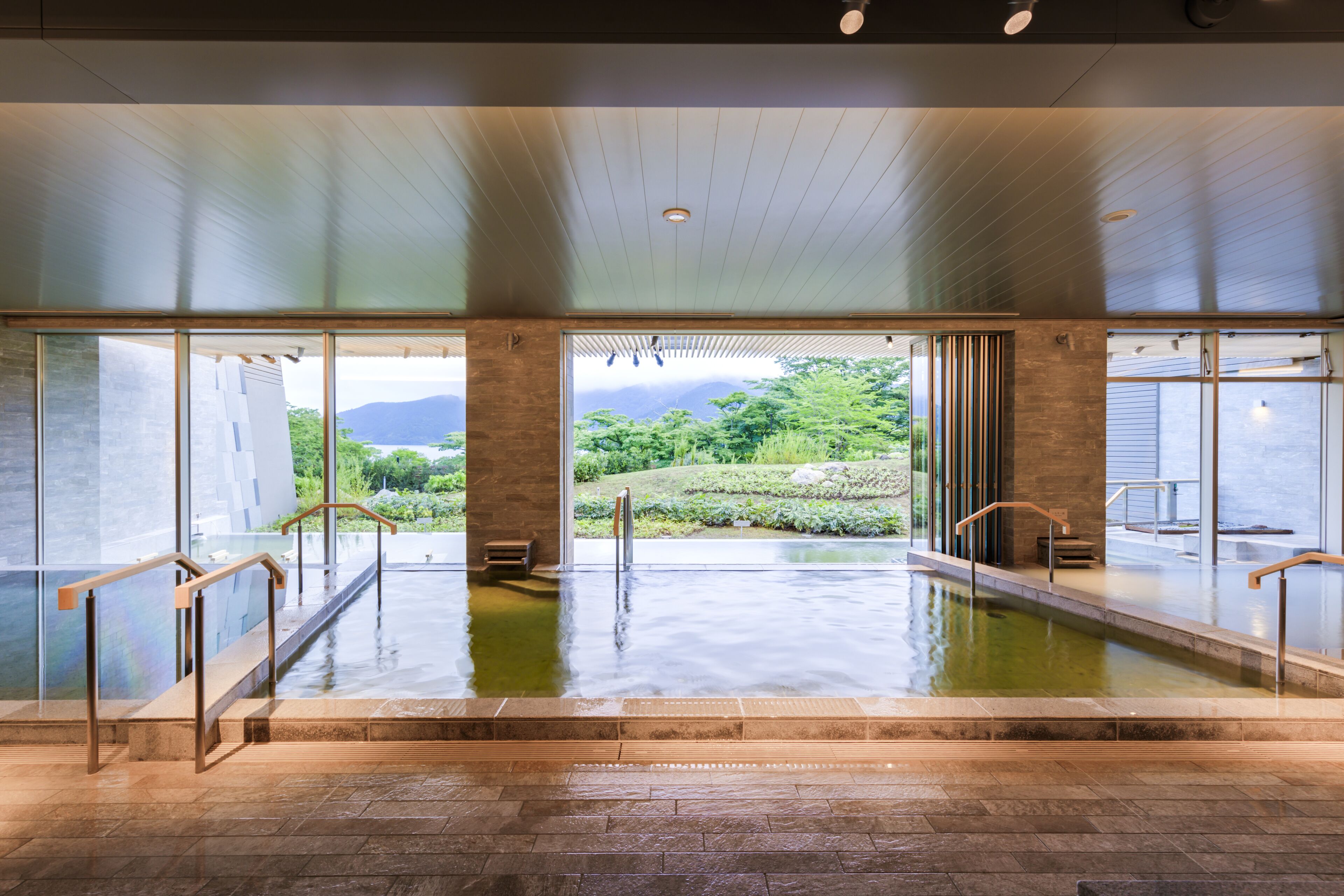 Piscina interior moderna com paredes de vidro transparente, guarda-corpos de madeira e vista para vegetação exuberante e montanhas.