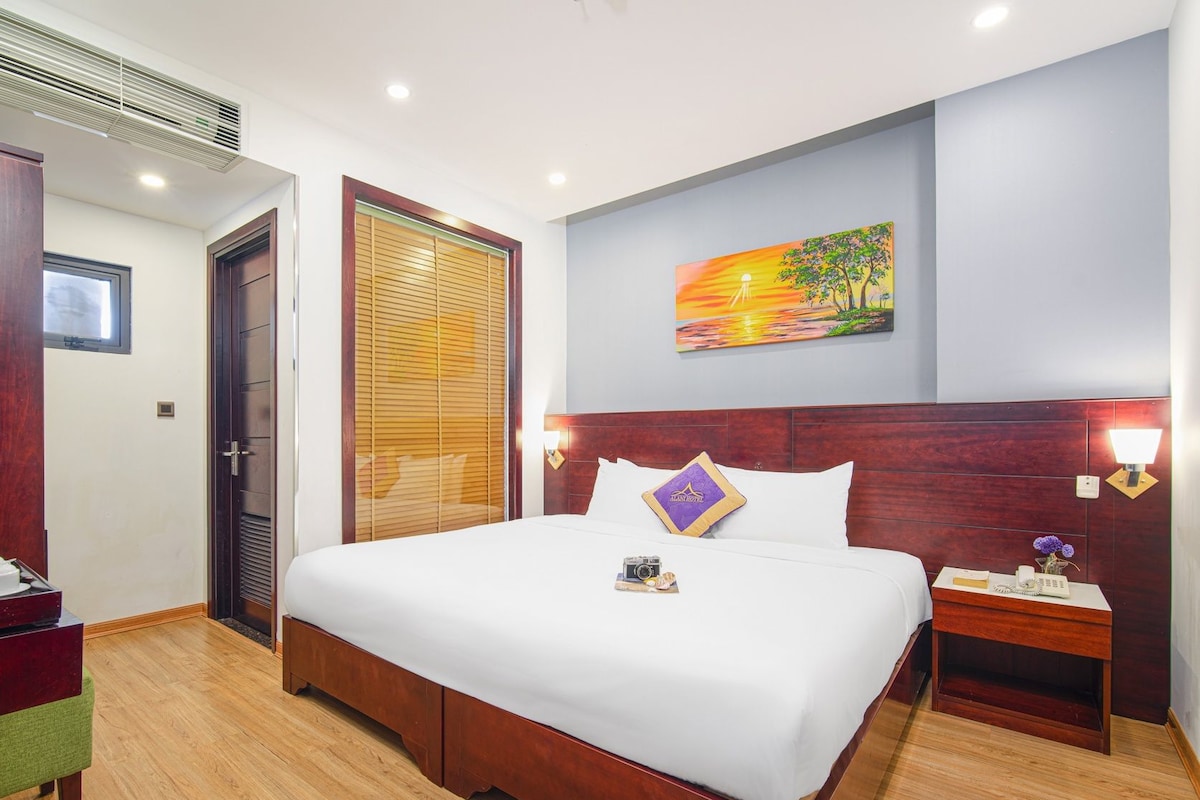 A hotel room with a large bed, a nightstand, a wall-mounted lamp, and a painting of a sunset.
