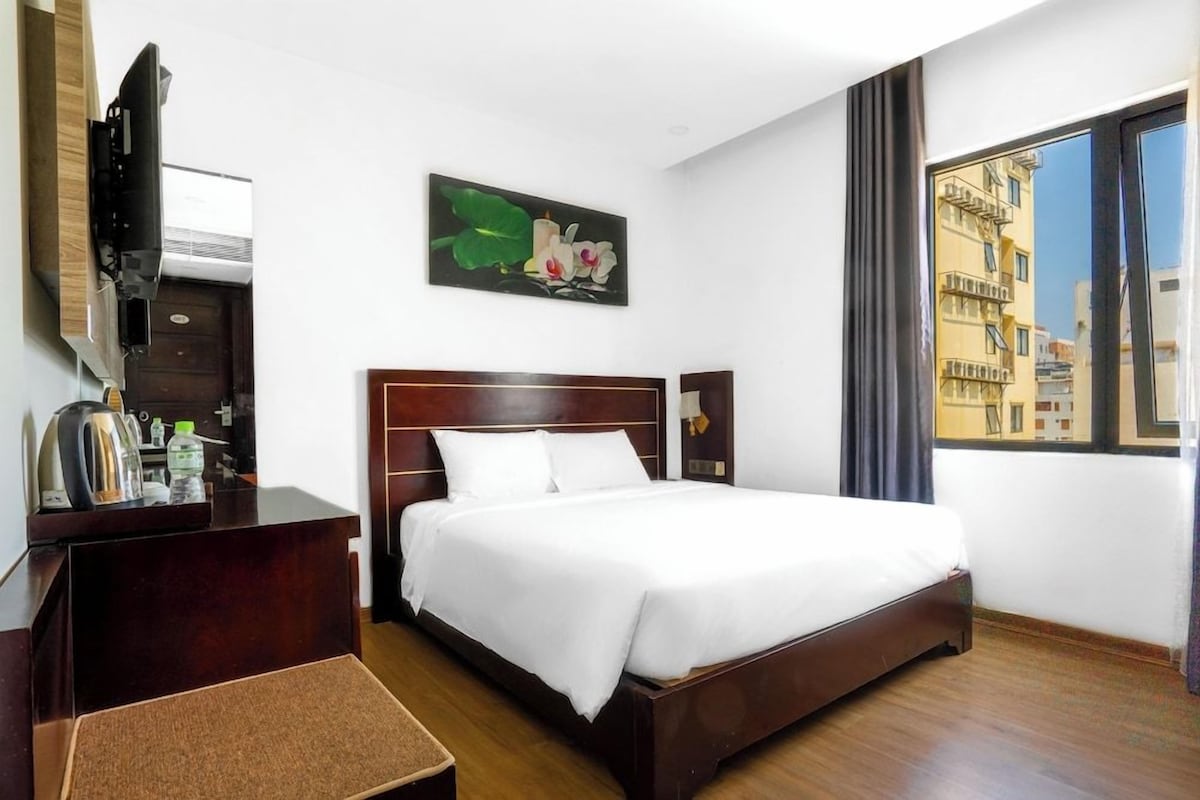 A hotel room with a bed, a desk with a television, a painting, and a window with a view of buildings.