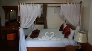 Un lit bien fait, avec du linge de lit blanc, un baldaquin et deux serviettes en forme de cygne. On y trouve une table de chevet en bois avec une lampe, un bureau en bois avec un miroir et une boîte de rangement orange.