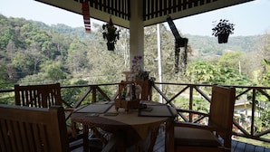 Une terrasse en bois avec une table et des chaises, entourée de plantes suspendues et offrant une vue sur une colline boisée.
